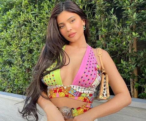 A woman with long dark hair wearing a colorful dress poses outdoors near green foliage.