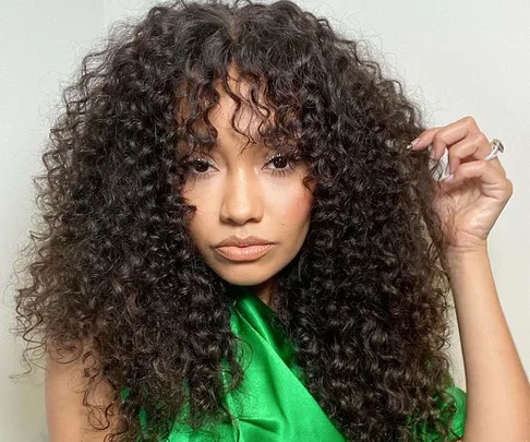 A woman with long, curly hair and a green top stands against a plain background.