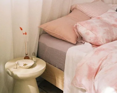 Cozy bedroom with pink and white bedding, bedside table, and decorative items in soft lighting.