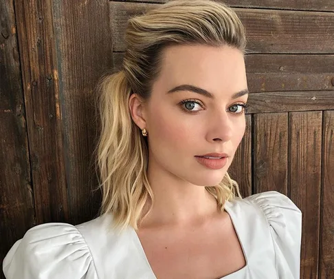 Blonde woman with styled hair against a wooden background, wearing a white top and gold earrings.