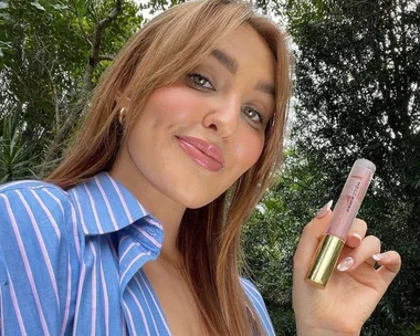 Person holding a Mecca Max lip product, smiling outdoors in a striped shirt.