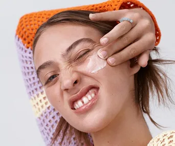 A person applying cream to their face with a playful expression, wearing an orange and purple knitted top.