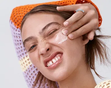 A person applying cream to their face with a playful expression, wearing an orange and purple knitted top.