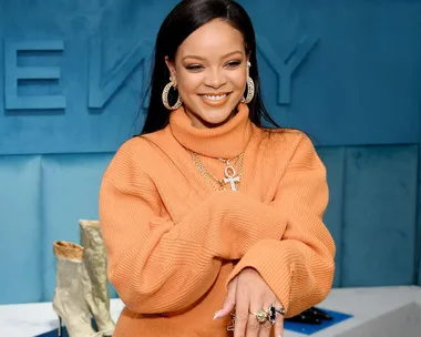 Rihanna wearing an orange sweater, smiling, with jewelry, at a Fenty event.