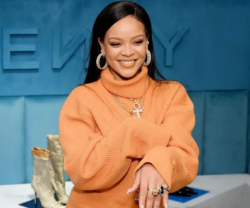 Rihanna wearing an orange sweater, smiling, with jewelry, at a Fenty event.