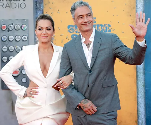 Rita Ora and Taika Waititi in formal attire pose together on the red carpet, with Taika waving and smiling.