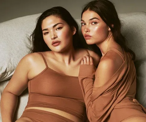 Two women in SKIMS lounge wear, seated closely, posing for a fashion shoot with neutral expressions.