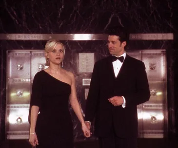 A woman in a black dress and a man in a tuxedo hold hands, standing in front of a marble wall.