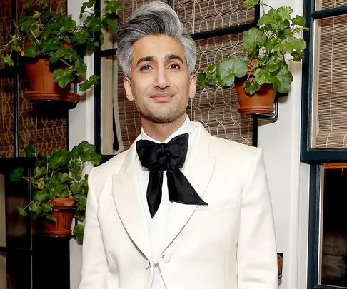 Person in white suit and black bowtie smiling, standing indoors with potted plants on shelves in the background.