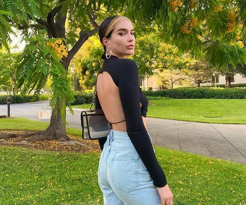 Woman with a tie-back black top and jeans stands in a park, surrounded by lush green trees and flowering branches.