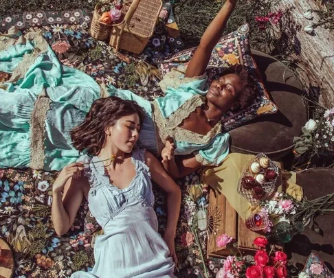 Two women relax on a floral picnic rug with snacks, a wicker basket, and flowers in a sunny outdoor setting.