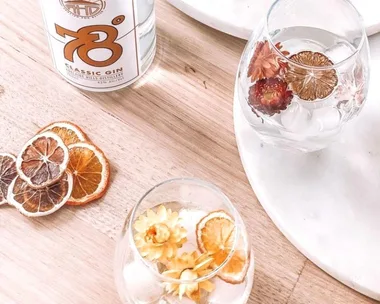 Two glasses of gin with dried citrus and flowers on a wooden table beside a bottle of 78° Classic Gin.