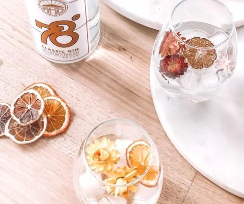 Two glasses of gin with dried citrus and flowers on a wooden table beside a bottle of 78° Classic Gin.