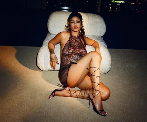 Woman in black lace-up heels and shimmery dress, sitting on a modern white chair, posing confidently.