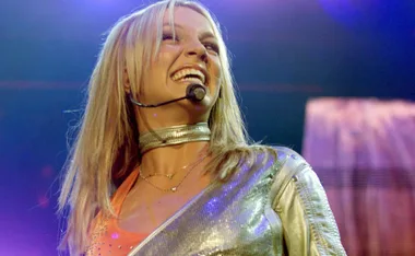 Singer performing on stage, wearing a silver outfit and headset microphone, smiling brightly under concert lights.