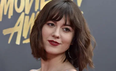 A woman with short brown hair and red lipstick posing on a red carpet in front of a "Movie Awards" backdrop.