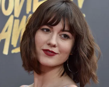 A woman with short brown hair and red lipstick posing on a red carpet in front of a "Movie Awards" backdrop.