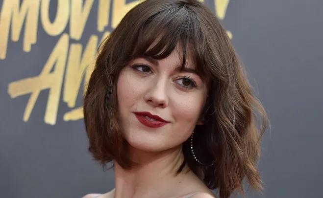 A woman with short brown hair and red lipstick posing on a red carpet in front of a "Movie Awards" backdrop.