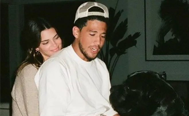 A woman hugs a man from behind while they both look at a black dog indoors.