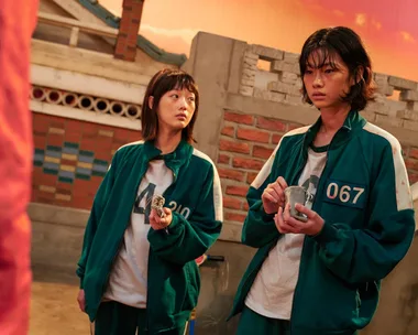 Two "Squid Game" characters in green tracksuits hold small bags in a brick courtyard, with a sunset sky in the background.