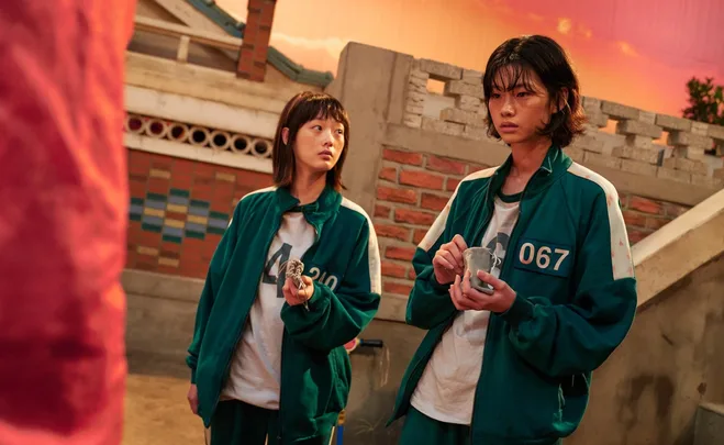 Two "Squid Game" characters in green tracksuits hold small bags in a brick courtyard, with a sunset sky in the background.