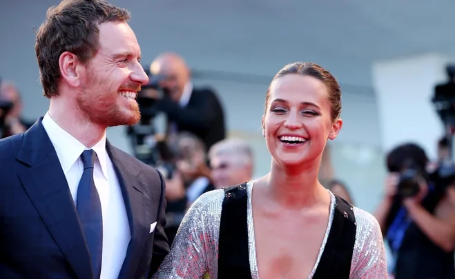 Smiling couple on a red carpet, he in a dark suit and tie, she in a sparkling dress, with photographers in the background.