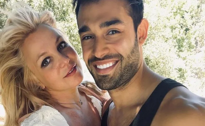 A woman with long blonde hair and a man with a beard smile at the camera, surrounded by green foliage.