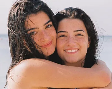 Two smiling young women with wet hair at the beach embrace each other against a blurry ocean background.