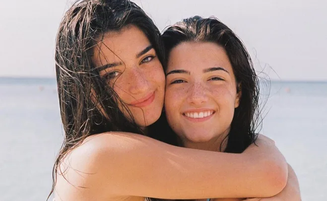 Two smiling young women with wet hair at the beach embrace each other against a blurry ocean background.