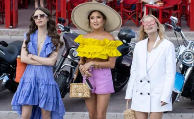 Three women from "Emily in Paris" in colorful outfits pose stylishly in front of motorcycles.