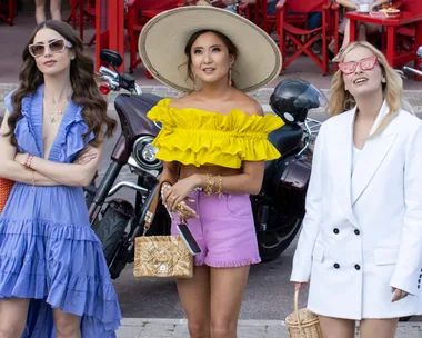 Three women from "Emily in Paris" in colorful outfits pose stylishly in front of motorcycles.