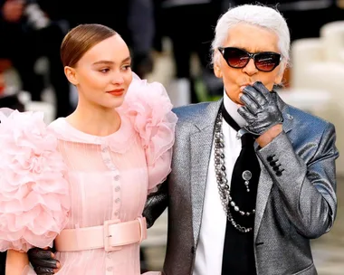 Fashion duo at event: woman in pink dress with ruffled sleeves, and older man in silver suit wearing sunglasses.