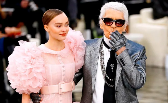 Fashion duo at event: woman in pink dress with ruffled sleeves, and older man in silver suit wearing sunglasses.