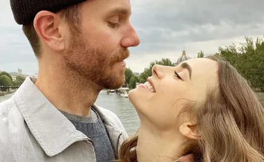 A couple smiling and looking at each other by a river on a cloudy day.