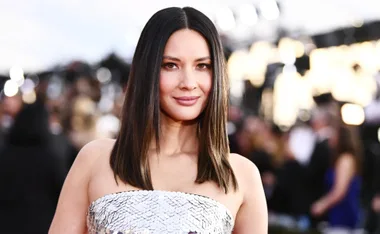 Olivia Munn in a strapless silver dress at a formal event, with blurred background lights.