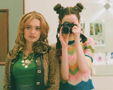 Two young women in a bathroom, one with a camera, the other in a green top and leopard jacket.