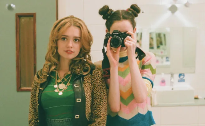 Two young women in a bathroom, one with a camera, the other in a green top and leopard jacket.