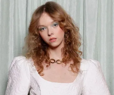 Young person with wavy hair in a white dress and gold necklace, standing against a light curtain backdrop.