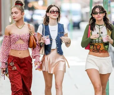 Three fashionable women walk confidently, wearing trendy outfits and holding drinks on a city street during fashion week.