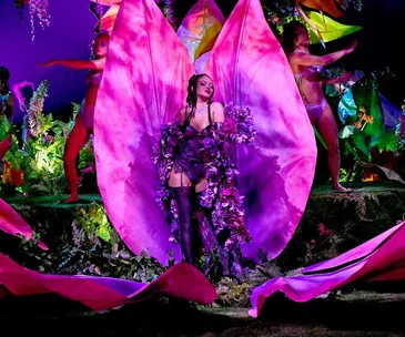 Vibrant fashion show scene with a model in purple floral attire, standing in large pink flower petals, surrounded by performers.