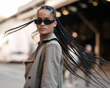 Person with long braids and sunglasses in a trench coat, walking confidently in an urban setting.