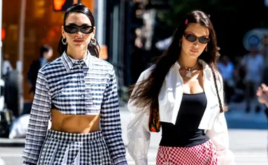 Two women walking on a sunny street, one in a plaid outfit, the other in a white top and red skirt, wearing sunglasses.