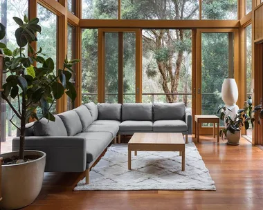 Spacious lounge with large windows, grey sofa, wooden table, and indoor plants overlooking lush greenery.