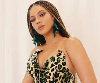 A woman in a leopard print dress with braided hair and green flower earrings, standing against a white background.