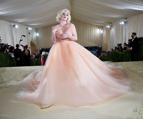 Billie Eilish in a flowing pale peach gown at the 2021 Met Gala, posing on the red carpet under a draped ceiling.