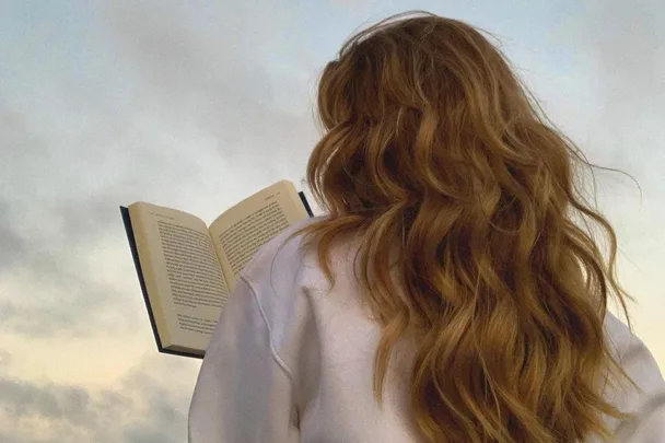 A person with long, wavy hair reads a book against a cloudy sky backdrop.