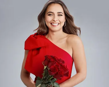 Smiling woman in a red dress holding a bouquet of red roses against a plain background.