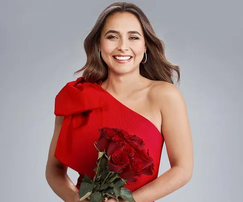 Smiling woman in a red dress holding a bouquet of red roses against a plain background.