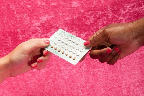 Two hands holding a pack of birth control pills against a pink background.