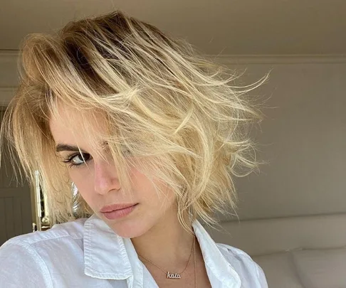 A person with tousled, blonde hair and a thoughtful expression, wearing a white shirt.